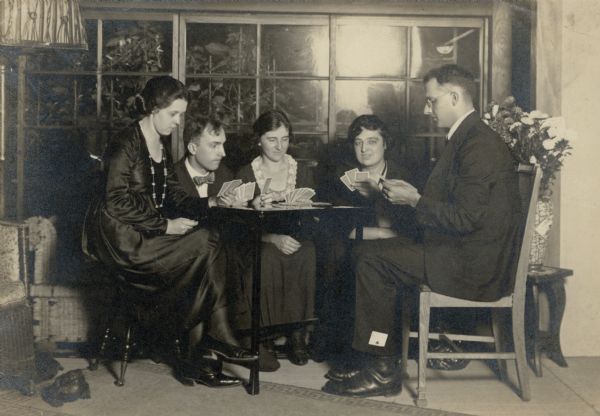 Leonore and Forest Middleton, on the far left, playing cards with two other women and a man while sitting at a small table. The man on the far right has the ace of clubs in his pants cuff. There is a pottery frog on the floor at left, and a vase with flowers on a small table at far right. The players are all well-dressed. On the reverse of the print Leonore has written: "Sept. 1921 Tokyo, Japan. Note the beads — Japanese crystal — the loveliest beads F. bot [sic] for me this summer. Taken in the Japanese house we lived in this summer. Just a friendly game — just posed — (flowers are asters). The man & lady in the center is Dr. Norden [?] & wife & the other girl is Miss Shoemacher who roomed with me this summer. She is a stenographer for the national [unreadable]." 