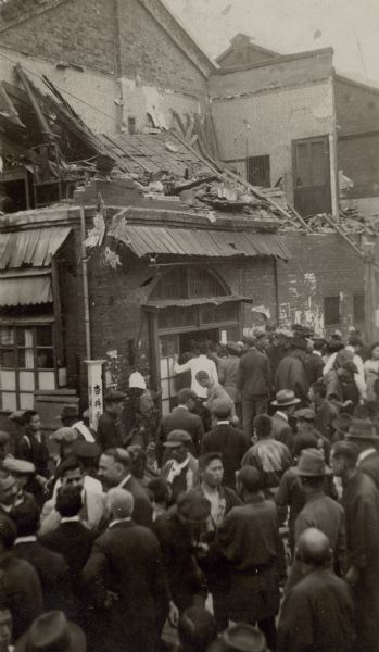 Elevated view of a crowd of men, some in traditional Japanese dress, and some in western-style clothing, gathered around a damaged building on a corner. The roof and second story walls of the small commercial building have collapsed. There is a signpost with Japanese lettering near the corner of the building. On the reverse of the print is written: "Pictures of the work of the earthquake that took place in Yokohama April 26, 1922."    