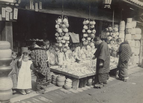 A shopkeeper is using a feather duster to clean stacks of bowls displayed on a low table in the open air. Teapots in an array of shapes and finishes are suspended from wires supported by bamboo poles. There are three children on the left; the oldest child is carrying an infant on his back. A woman at right is looking over the merchandise. There are large pots stacked at far right.  