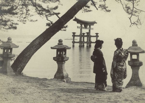 Stone lanterns on the shore near a leaning pine tree frame a view of the Great Torii on Japan's Inland Sea. Two Japanese women in traditional kimonos are standing on the shore, facing the gate. This photograph was taken near the Itsukushima Shrine on Miyajima Island.  