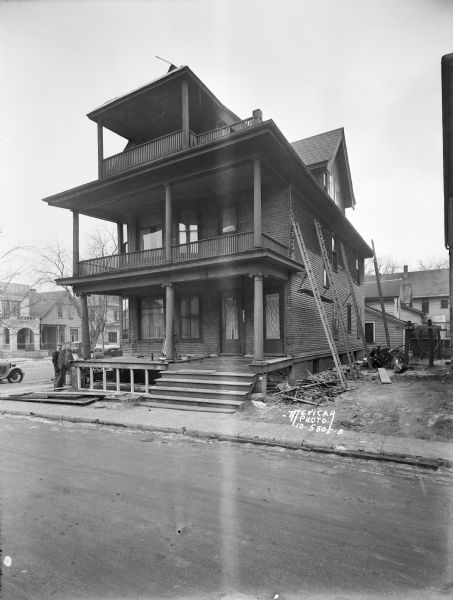 View across street towards a three-story house on Bernard Court. The address on the door reads "202," and a sled is leaning up against the wall near the front windows. There are ladders set up against the right side of the house for working on the siding by the Niesen Building Co. Two men are standing on the left near the front porch.