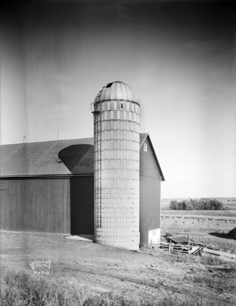 View of dome roof silo with a small door at the top open, built by Madison Silo Co., 3222 Atwood Avenue, standing next to a barn.