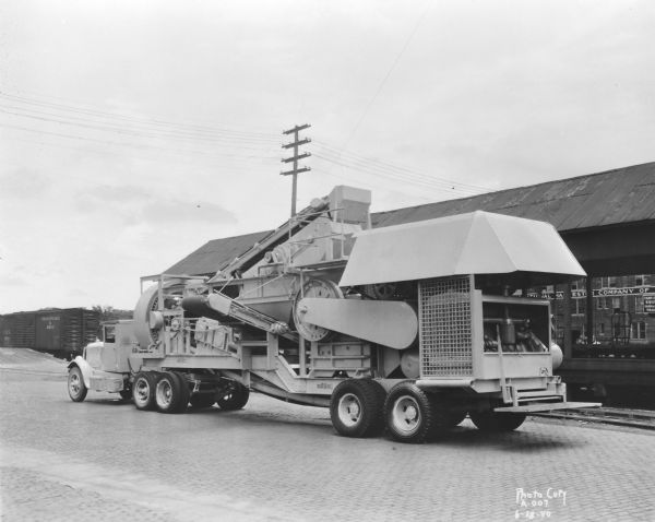 Truck-pulled rock crusher manufactured by Wisconsin Foundry & Machine Company, 623 East Main Street. Left rear view of the unit parked along railroad tracks. In the background is the International Harvester Company of America building, 301 South Blount Street, and a Chesapeake & Ohio Railroad boxcar.