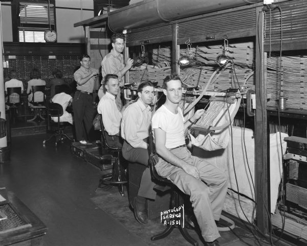 Five men are rebuilding the "Fairchild" exchange switchboard at the Wisconsin Telephone Company building. Four women telephone operators in the background are operating another exchange switchboard.