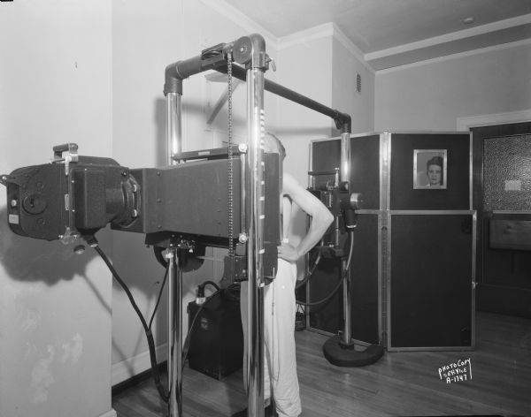 State of Wisconsin General Hospital, at 1300 University Avenue. View showing chest X-ray equipment with a patient in position for the procedure, and a technician behind the protective screen.
