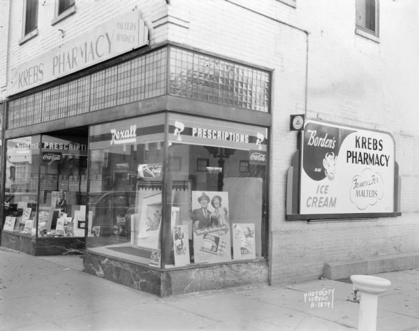 View towards the Kreb's Pharmacy on a street corner. There are Rexall and Borden's Ice Cream signs in the window. There is a fountain (bubbler) on the sidewalk near the curb.