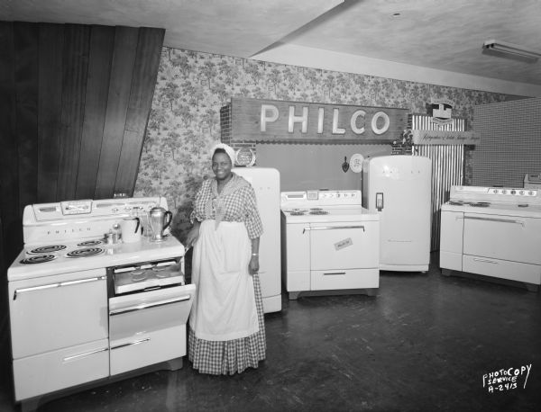 A woman dressed as Aunt Jemima is making pancakes on a pull-out griddle at a stove which is part of a merchandise display at Evans Radio and TV Store, 4233 West Beltline Highway, featuring Philco appliances including two refrigerators and three stoves.
