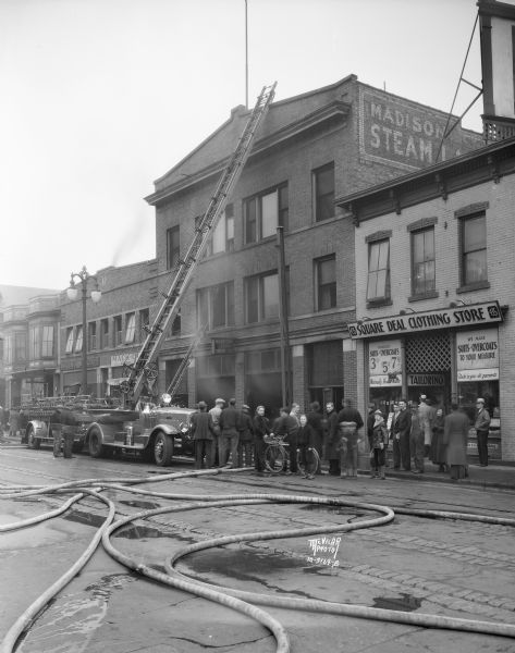 View across street towards the Madison Steam Laundry, at 433 State Street, the scene of a fire. In front of the building is a fire truck with extension ladder. Fire hoses are laying in the street, and spectators are standing and watching from in front of the Square Deal Clothing Store, at 435 State Street.