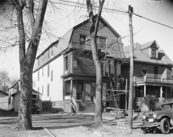 View from street of the workers re-siding a house at 412 W. Mifflin Street. An automobile is parked in front of the garage on the left, and there is a neighboring house on the right. Two men are standing on scaffolding on the right side of the building.