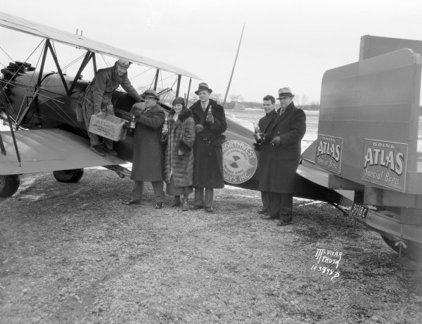 Ira Chambers of the Atlas Distributing Co. is shown receiving the first case of Atlas Beer, of Chicago, Illinois, from the pilot of a Northwest Airlines plane at the airport. Three other men and one woman are standing nearby holding bottles of beer.