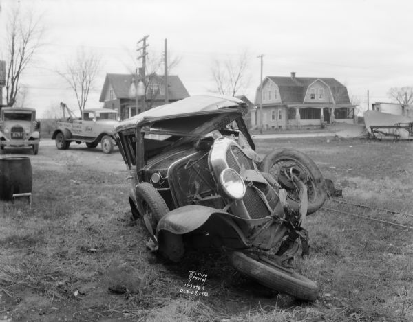 View from front of a wrecked Oakland Sedan. There is a tow truck and houses in the background.