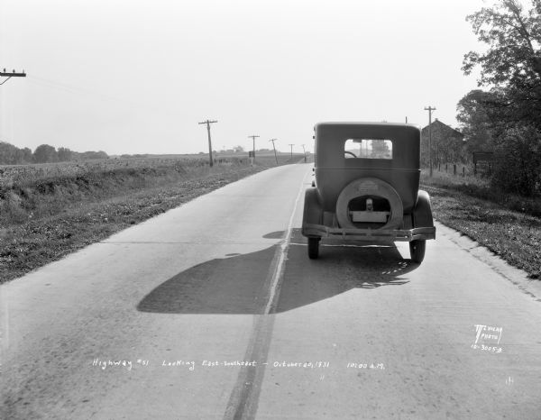 View looking down road (east-south east) towards the rear of a car parked in the right lane of a two lane highway. This was the scene of an accident, and is showing the visibility of a car on Highway 51. A man is standing on the side of the road on the left in the distance.