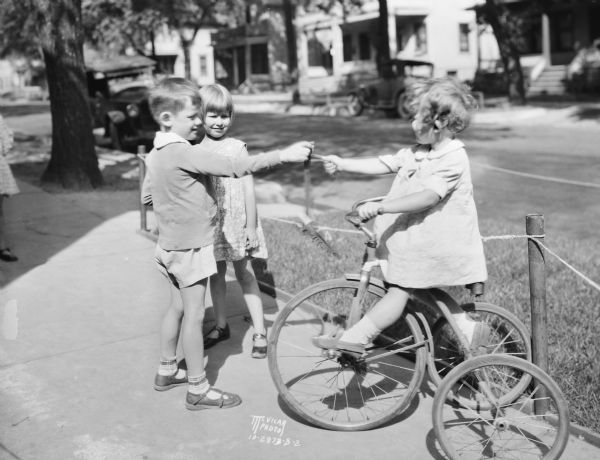 Two school children are playing with a girl sitting on a tricycle while walking to school.