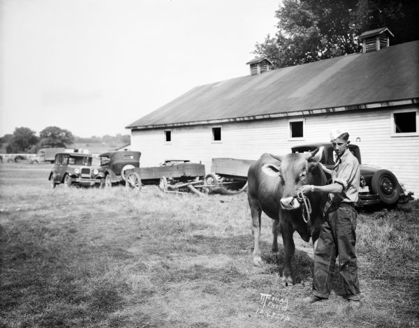 Mr. Olson is standing outdoors with a Brown Swiss bull at the Dane County fair. Automobiles and wagons are parked along a wooden building in the background.