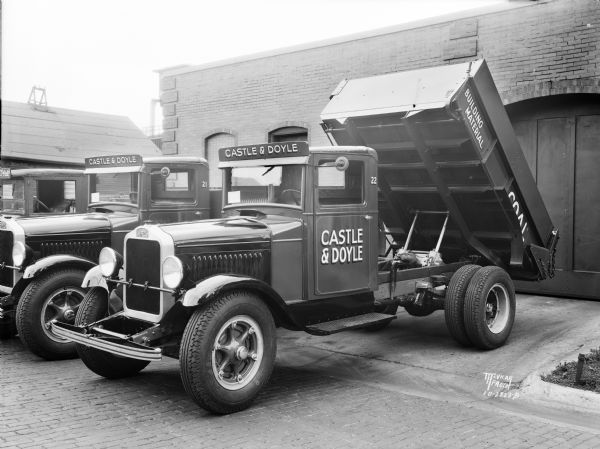 White Motor Company Castle & Doyle truck, with the bed of the truck in the dumping position, backed up to a building. The signs painted on the bed of the truck read: "Building Material" and "Coal." Two more trucks are parked on the left.