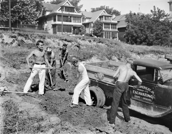 Five University of Wisconsin-Madison athletes (L. to R. Walter Gnabah, Russel Rebholz, George Wright, McClure Thompson, and William Eller) shoveling dirt at Camp Randall with a Fay Hammersley, Jr. Excavating truck parked nearby. Houses on Breese Terrace are in the background.