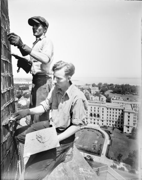 Close-up of Don Coffey, in the foreground, and Bruce Russell, wearing the cap, steeplejacks repointing the University of Wisconsin heating plant chimney. Photograph taken by Aldro Wasley on the scaffold 192 feet in the air. In the background is an elevated view of the U.W. Hospital, Elizabeth Waters and Home Economics building. In the far background is Lake Mendota and Picnic Point.