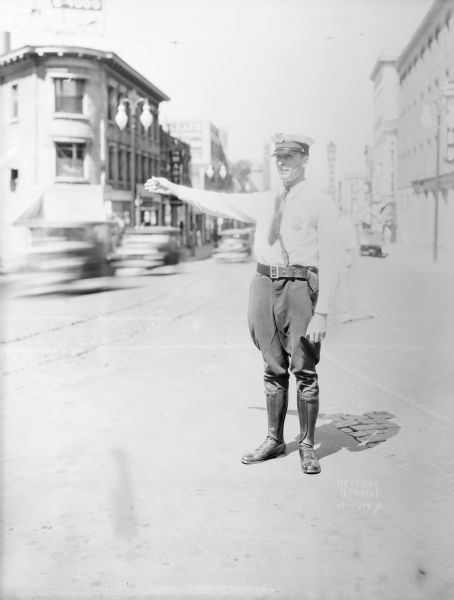 View of a policeman standing in the street and directing traffic on State Street. Moving cars are in the background.