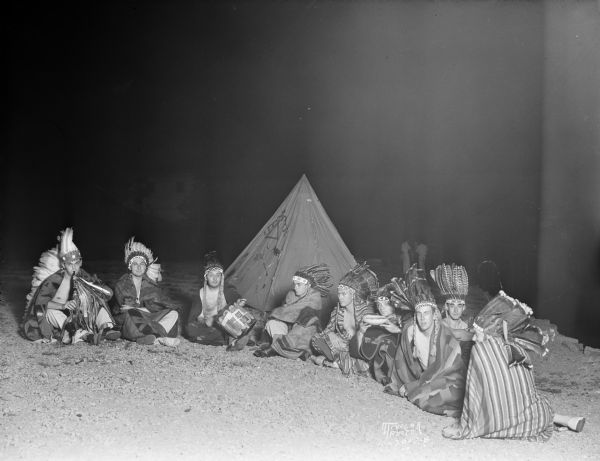Nine members of the junior and senior class at the University of Wisconsin are sitting in a semi-circle dressed as Native Americans. They are smoking the pipe of peace on the Mendota lakeshore. A tent is in the background.