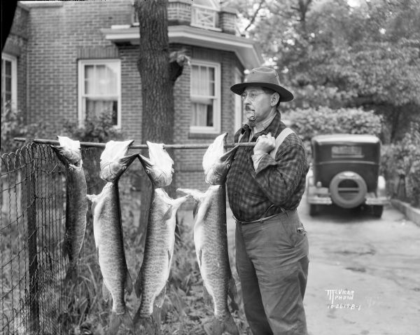 Dr. Karl Smith is standing near his house at 1 N. Prospect Avenue. He is holding one end of a pole that is resting on a fence and displaying four muskellunge strung on it. He caught the fish on the Larson islands in Vilas county.