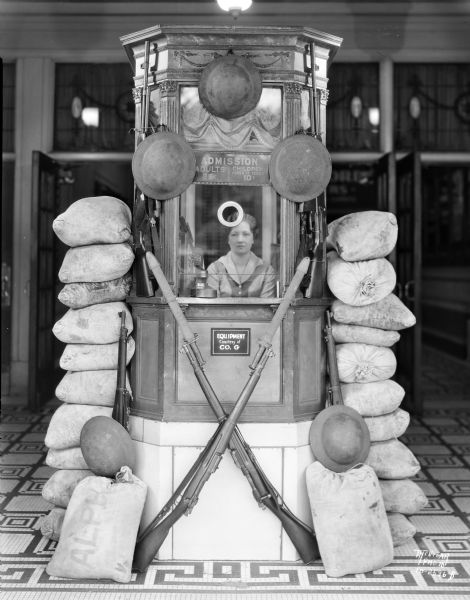 View towards a woman sitting in the Strand Theatre box office surrounded by a display of helmets, guns and sand bags for the motion picture "Women of All Nations," the third movie of Edmund Lowe and Victor McLaglen acting as marines.