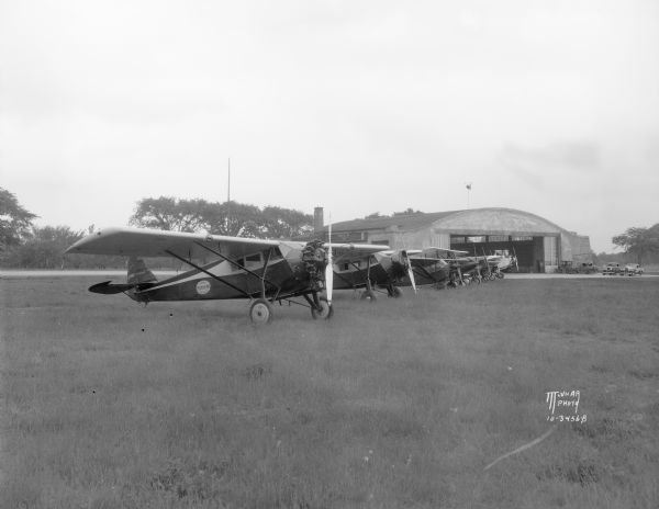 At Royal Airport, a line of airplanes are parked in a row in a field. In the background is a hangar with automobiles parked nearby.