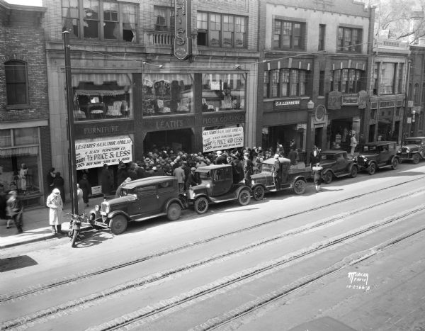 Elevated view looking across street towards a crowd waiting to attend the opening of the Black Furniture Company's bankrupt sale at Leath's furniture store, at 117-119 State Street. Storefronts nearby are the G.A. Lehnherr's ladies' wear, at 121 State Street, Glasgow Tailors, at 123 State Street, and Castle & Doyle Coal Co., 125 State Street. There is a row of automobiles and a motorcycle parked at the curb.