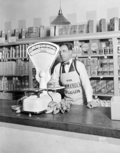 Interior view of a Toledo Computogram scale on a counter in a Dodgeville store, with the grocer weighing bananas. In the background is a display of merchandise on shelves.