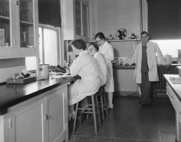 Four laboratory workers in the Madison General Hospital laboratory.  Three women are working at a counter near microscopes. A man is leaning against a counter in the background.