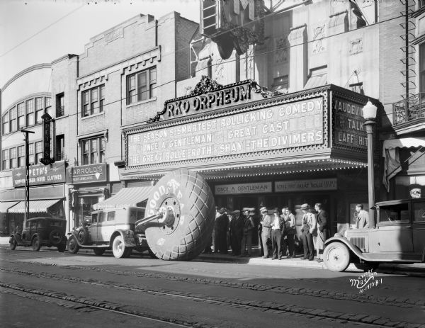 View across street towards the World's largest tire on display in front of the RKO Orpheum Theatre at 216 State Street. A group of people are standing on the sidewalk looking at the tire which is attached to a truck. The theater marquee lists a movie: "Once a Gentleman." Two storefronts on the right are Harrold's Credit Jewelers and Speth's Clothing Store.