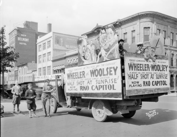 Two men and two boys are standing besides a truck parked on North Pinckney Street which has large signs advertising Bert Wheeler and Robert Woolsey in the movie "Half Shot at Sunrise." Two people are standing in the bed of the truck. In the background is the New Belmont Hotel and Harry Manchester.