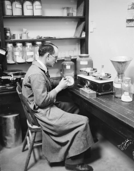 View of a man taking Sodium Chloride (NaCl) out of a jar to measure it on an old scale on a table. The man is working in the Ricketts laboratory at the Agriculture Chemistry Building at the University of Wisconsin-Madison.