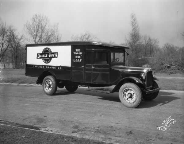 View towards a Gardner Baking Co. truck with "Gardner's Double-Duty Bread" and "The 'TWO IN ONE' Loaf" signs on the side. The truck is parked in the center of a street.
