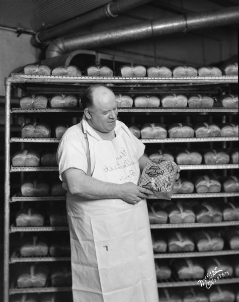 A baker is standing and holding a loaf of Gardner's Double Duty bread, with shelves of bread in the background. The man is wearing Else Russell's Best Flour apron.