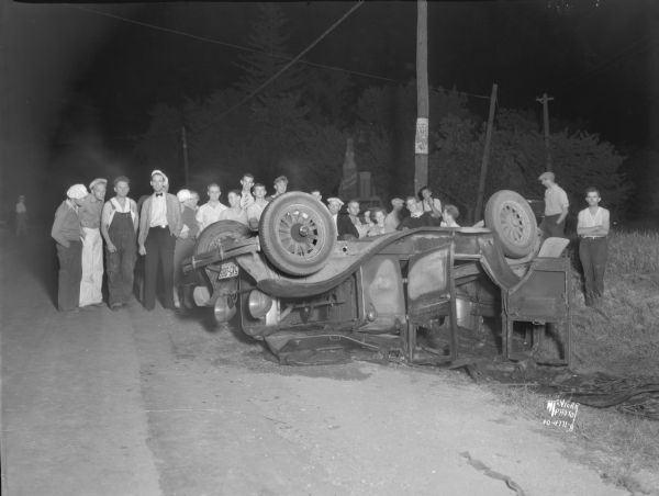View down road towards a group of people looking at an overturned automobile belonging to Fred Swanson. Fred and Norma Swanson, Dorothy Thompson, Edward and Belva Hotmar, and George and Bernice Radke were hurt in a collision at the intersection of East Washington Avenue (Sun Prairie Road) and Fair Oaks Avenue. The Hotmar car struck the Swanson car in the rear, overturning the Swanson car which immediately burst into flames.