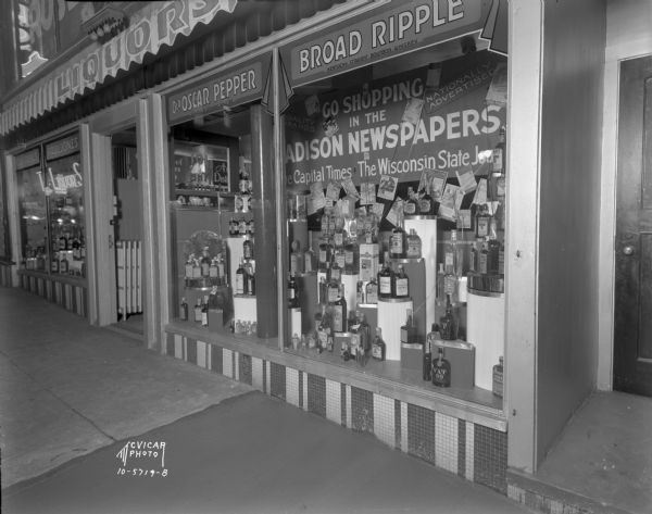 The Liquor Shop, at 101 West Mifflin Street. Liquor bottles are on display in the show windows. A sign in the window reads: "Go Shopping in the Madison Newspapers, The Capital Times, The Wisconsin State Journal."