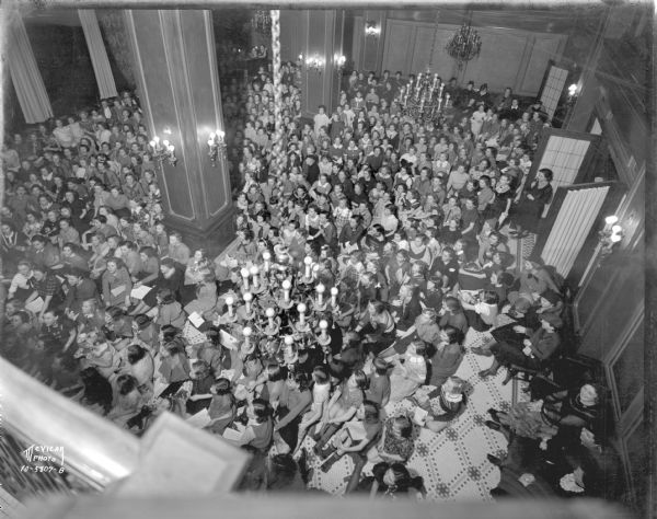 Elevated view from balcony of large group of Girl Scouts in the dining room of the Loraine Hotel. Many of the girls are sitting on the floor in the front, with other girls and woman sitting and standing near the back of the room.