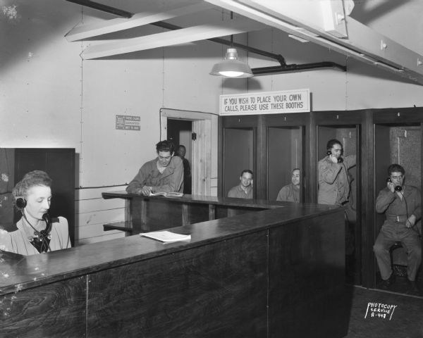 View of a group of men and a woman. Some of the men are dressed in overalls and are using telephones in a bank of telephone booths at Truax Army Air Field. A woman wearing a telephone headset is sitting behind a counter on the left. This image was made for the Wisconsin Telephone Company.