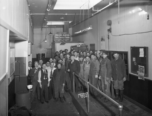 Group portrait of Oscar Mayer stockyard employees, and one man in a business suit, gathered near the Farm Service Studio in the Oscar Mayer plant, 910 Mayer Avenue. Overhead sign reads: "On the Air, Oscar Mayer Farm Service Studio, WIBA, WIBU."