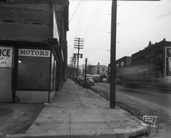 View down sidewalk along East Wilson Street at the corner of the South Hancock intersection looking toward the West Wilson Street-King Street intersection. The photograph was taken to document the site of a Reynolds Bus accident scene. The business seen at the near corner are Electric Motor Service at 323 East Wilson Street. The business seen at the far corner is Schroeder & Son Funeral Service, at 235 King Street.  Businesses along the north side of East Wilson Street include the Shell Service at 324 East Wilson Street, R & R Cafe at 316 East Wilson Street, Madison Watch Shop at 314 East Wilson Street, Capitol City Furniture Co. at 310-12 East Wilson Street, Fred Kessenich (United Autographic Register Company) at 308 East Wilson Street, Johnson Maytag Company at 306 East Wilson Street, Meyer Printing at 304 East Wilson Street, and Stop Lite Tavern, 302 East Wilson Street.