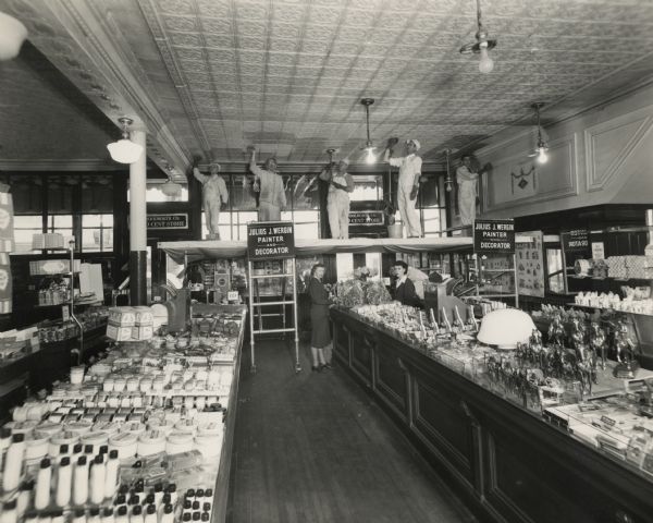 Elevated view of two sales women of the F.W. Woolworth Co. standing at a counter, while five men, from Julius J. Wergin Co. painters and decorators, are standing on scaffolding above them applying paint to a tin ceiling. Long wooden counters display merchandise, including rows of horse statues of different sizes. Wood flooring is seen between the counters. In the background, daylight is shining through the upper portion of the store's front display windows. Woolworth Co. signs are above two double-door entrances. The F.W. Woolworth Co. was the successor of the F. M. Kirby Co.'s 5 and 10 Cent Store located at 907 S. 8th Street. In 1912, F.M. Kirby, who owned close to 100 stores in the U.S., joined Frank W. Woolworth to become part of F.W. Woolworth Co.'s chain of variety stores. In 1918, the store was expanded into the adjacent building at 905 S. 8th Street. The two buildings were joined and remodeled, which included a new storefront. The company continued in operation at 905-907 S. 8th until moving to the new Park Plaza Shopping Center in 1959. Julius J. Wergin's painting and decorating business was in operation from 1920-1970.