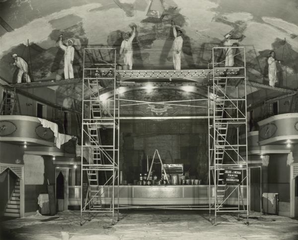 View of the Julius J. Wergin Company's decorating crew painting the ceiling while standing on scaffolding high above the New Opera House floor. Paint cans, a ladder and other supplies are on the stage. Drop cloths cover the floor. The stage and tiered box seats of the main ballroom are on the left and right. The Opera House was located at 411 N. 8th Street. Julius J. Wergin's business was in operation from 1920-1970.