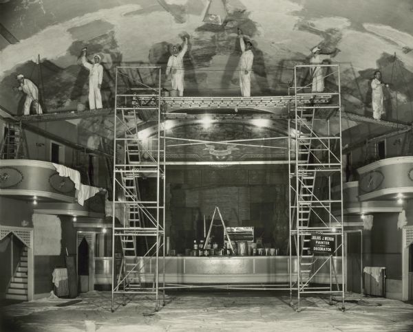 View of the Julius J. Wergin Company's decorating crew painting the ceiling while standing on scaffolding high above the New Opera House floor. Paint cans, a ladder and other supplies are on the stage. Drop cloths cover the floor. The stage and tiered box seats of the main ballroom are on the left and right.  The Opera House was located at 411 N. 8th Street.  Julius J. Wergin's business was in operation from 1920-1970. 