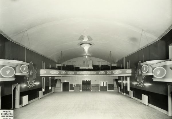 Elevated view of the rear of the New Opera House at 411 N. 8th Street following remodeling, with its ornately decorated walls, tiered boxes and projecting balcony. A large crystal chandelier, bracketed wall lighting and reflective, light-colored ceiling provide illumination. Open doorways, normally velvet draped, lead to upper-level seating. Steam-heat radiators line the walls. Painting and decorating by Julius J. Wergin.