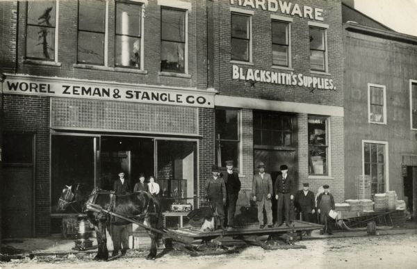 Albert Worel, Frank Zeman and Jacob Stangel are standing in front of the brick and wood-frame buildings of their York St. hardware store. The company's delivery horse, "Turk," is hitched up to take on a load. Standing at the front of the platform sleigh is the driver, Lewis J. Schroeder. Next in order on the sleigh are Frank Zeman, Elmer Dryer and Louis Wicke. Behind the sleigh, on the right, are Jacob J. Stangel and Frank Giese. In the doorway, behind the horse, are Albert Worel, Frank L. Stangel and Olivia Geisler. Located at 718-722 York Street, the buildings were connected to a storefront entrance at 205 N. 8th Street and operated under the ownership of the three partners from 1905-1917.  In 1905, Worel, Zeman and Stangel formed a partnership and purchased the hardware establishment of Rand & Roemer. The new firm carried on as ship chandlers supplying the marine trade, stocked blacksmith's, farmer's and hunter's supplies, shelfware, tinware, graniteware, as well as doing contracting and installation work. A Feb. 28, 1905 advertisement in the "Manitowoc Daily Herald" states: "Store Now open for Business, Worel, Zeman & Stangel Co., at the old Rand & Roemer Stand (Corner Eighth and York Streets.) Now established with complete, large and new stock of hardware, building material and everything necessary to a first class hardware store." In 1917, Jacob J. Stangel left to establish his own hardware business on S. 8th and Quay Streets, the J.J. Stangel Hardware Co.