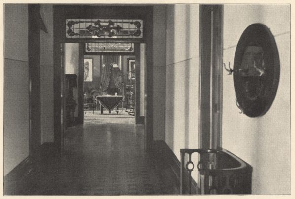 View inside the main entrance looking through the first floor corridor into a furnished room. A stained glass window with a floral motif is above the vestibule door. The stained glass window above the furnished room's entrance reads: "Parlor." A large piece of furniture, obscured by a wall, is in the corridor to the left of the parlor door. The vestibule has a door on each side, one with an open transom for air circulation. A mirrored hat rack and and umbrella stand are in the foreground on the right. The floor appears to be a wood parquet design. The first floor also had a doctor's office, apothecary, patient's sitting and dining rooms, furnished private rooms and a large ward for men.