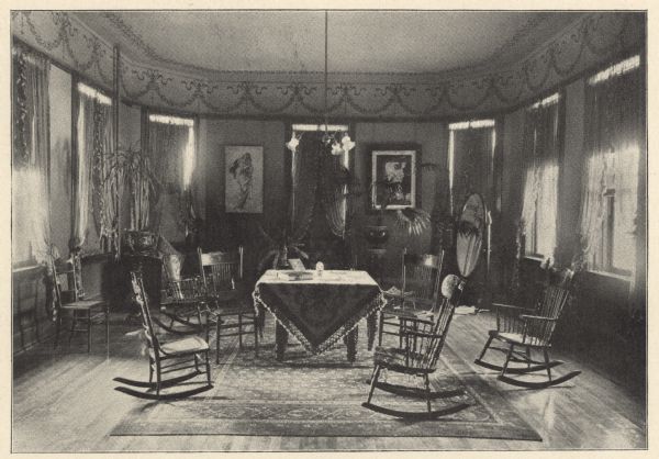 Elements of Victorian design and decoration are seen in the main floor parlor. Ribbon and floral wreathing draws attention to the upper walls and high ceiling. A wooden spiral turned-leg table with an ornately patterned and trimmed covering holds a book with a clasp; possibly a Bible. Wooden rocking chairs and side chairs have cane woven seats. A patterned rug partially covers a hardwood floor. Sheer ruffled-edge curtains are tied back just above the window sills. On the far wall, pictures are of Mary with Jesus on the left, and the Good Shepard with his sheep on the right. The diagonal corners of the room indicate its bay-shaped design, which extends from the west side of the building. The parlor was used for visiting patients. Friends were able to visit on Sundays, Tuesdays and Thursdays between the hours of 3:00 and 5:00 p.m.  Relatives were not restricted to the regular visiting days or hours.