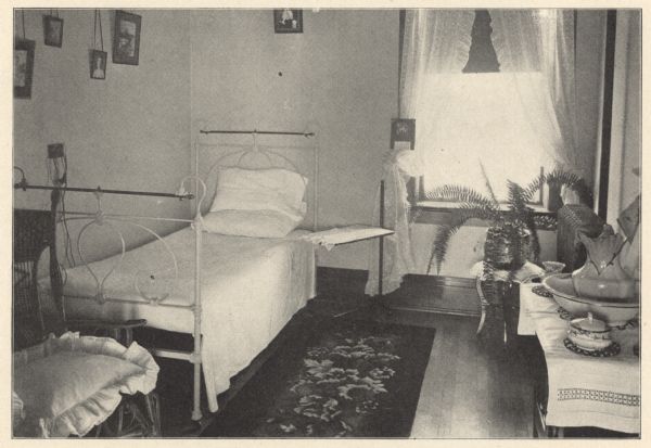 View of a small private room with a cast-iron and brass bed. A single window with sheer dotted Swiss tied-back curtains brightens the room. On the hardwood floor is a floral patterned rug. A wicker chair with plush seat cushion and cast-iron tray table are standing near the bed. On the right, a pitcher, basin and accessories are on a linen covered wash stand. High above the bed are framed pictures hanging from cording. The first floor was exclusively for male patients, while the second floor was reserved for female patients and a chapel. Private rooms ranged from $10 to $20 per week, depending on the location and character of the room.