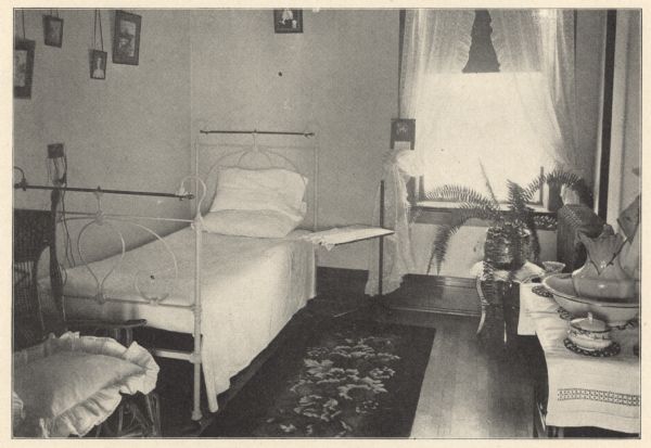 View of a small private room with a cast-iron and brass bed. A single window with sheer dotted Swiss tied-back curtains brightens the room.  On the hardwood floor is a floral patterned rug. A wicker chair with plush seat cushion and cast-iron tray table are standing near the bed. On the right, a pitcher, basin and accessories are on a linen covered wash stand. High above the bed are framed pictures hanging from cording. The first floor was exclusively for male patients, while the second floor was reserved for female patients and a chapel. Private rooms ranged from $10 to $20 per week, depending on the location and character of the room.