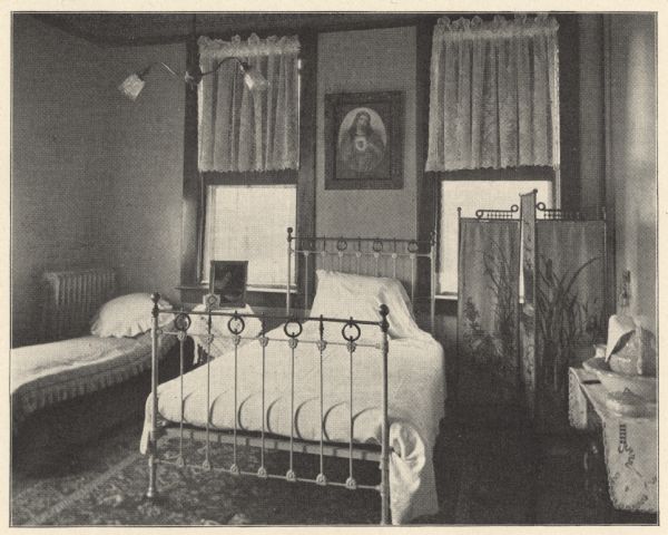 View of a large, private hospital room with a linen covered cast-iron and brass bed. Two windows with lace tier curtains allow light to enter the room, and a picture of the Sacred Heart of Mary is hanging on the wall between them. A daybed, steam heat radiator and bedside table holding a clock and framed picture are on the left. To the right is a three paneled folding privacy screen with a depiction of a nature scene. Along the wall is a washstand with covered pitcher and basin. A large patterned floor rug and double-shaded hanging light fixture complete the view. Private rooms ranged from $10 to $20 per week, depending on the location and character of the room.